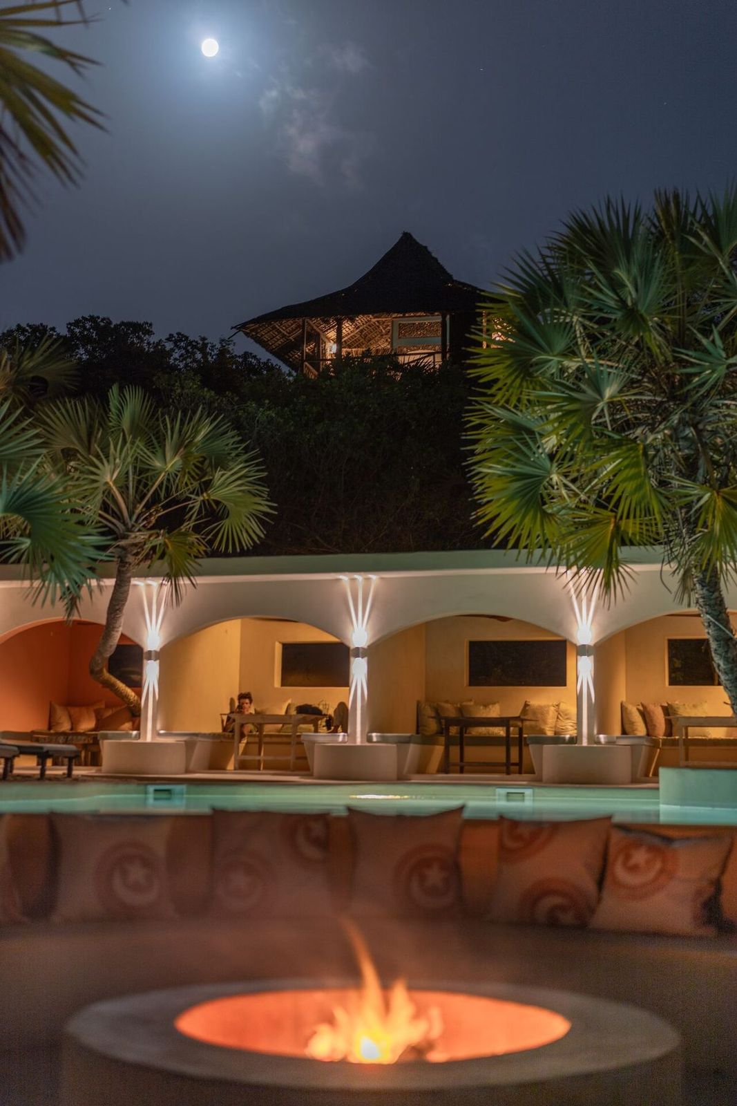 Firepit glowing beside the lit pool and arched colonnade at The Cabanas Lamu, full moon above the makuti rooftops