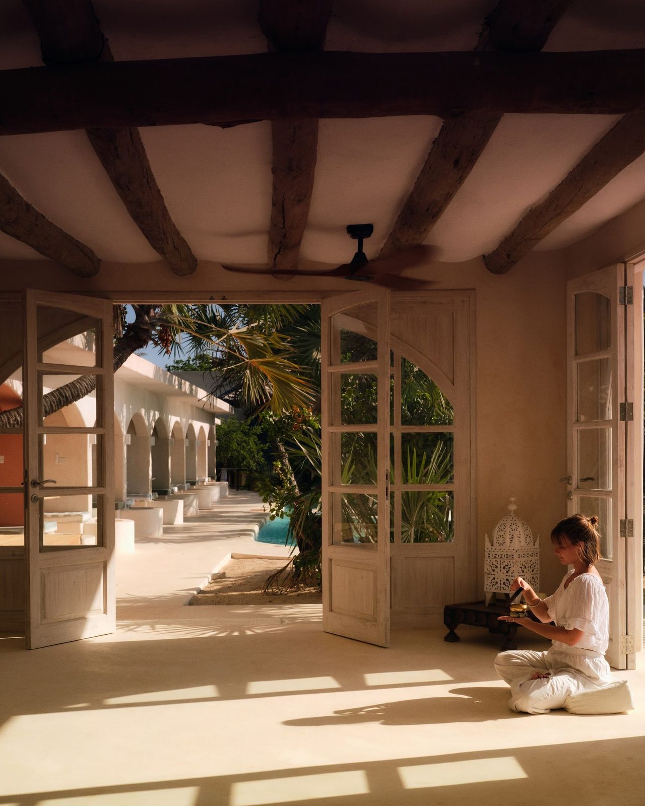 Yanna seated with a singing bowl in the Zen Den, the pool and palms beyond the open doors