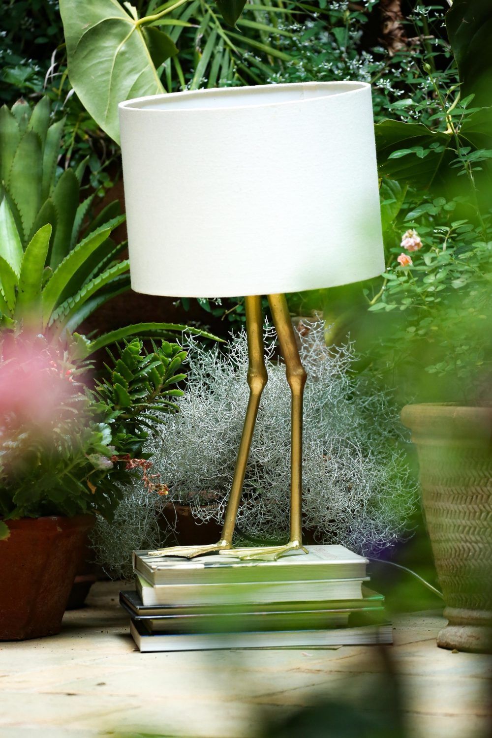 A brass crane-leg table lamp resting on a stack of books in a tropical garden