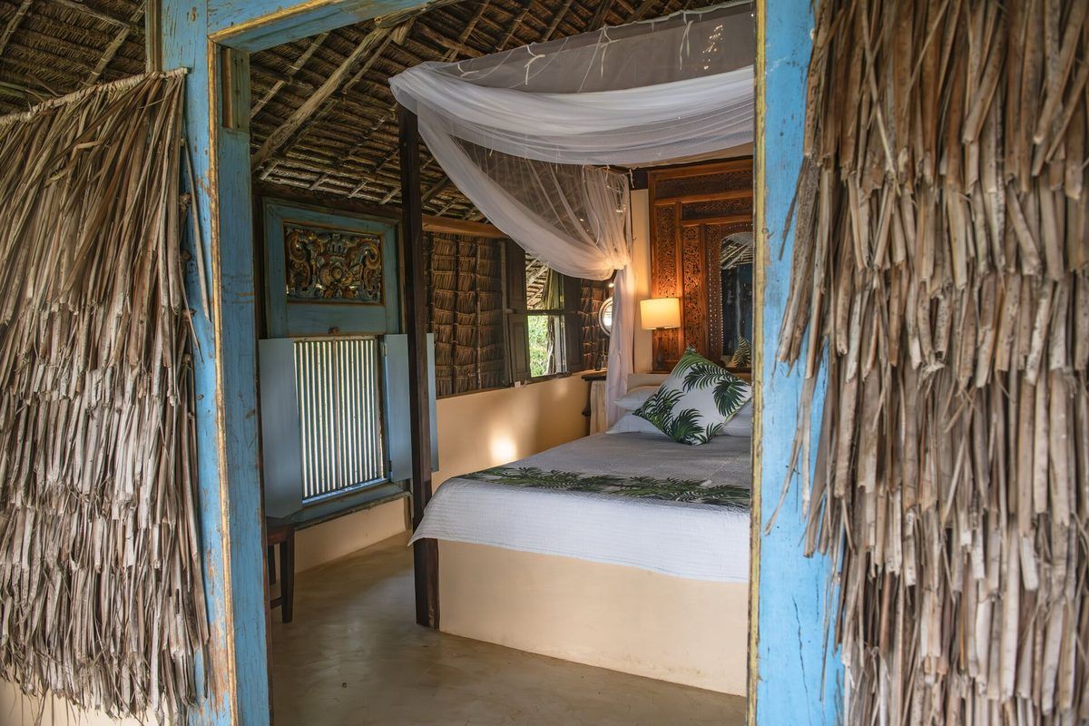 Moana cabana — double bed seen through a palm-thatch wall framed by a blue Bali doorway